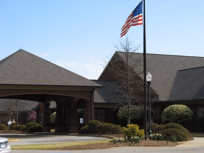 Brick retirement community entrance with a covered porte-cochère, American flag on a pole, and landscaped shrubs.