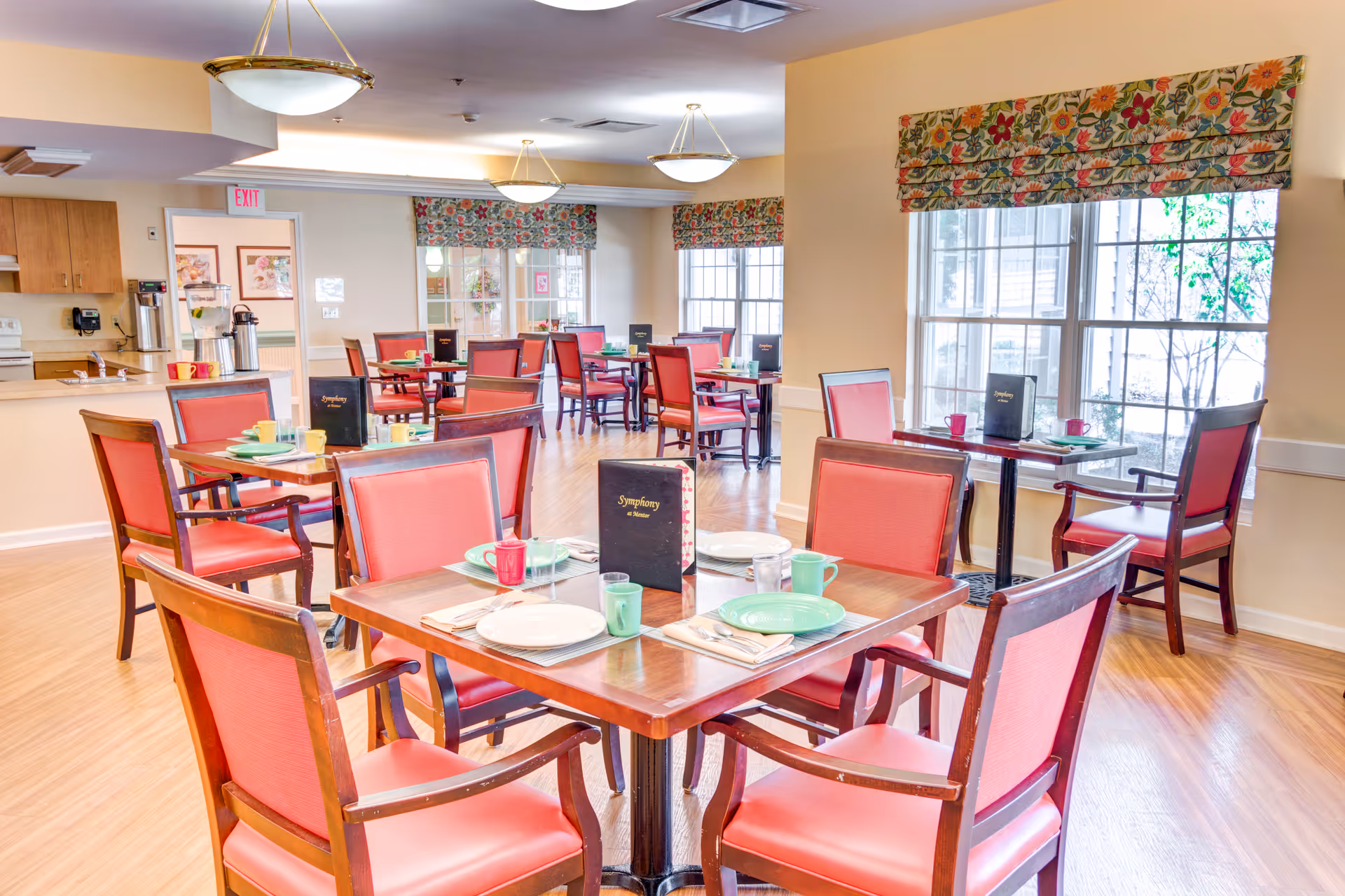 A bright dining room with wooden tables and red cushioned chairs. Each table is set with plates, cups, and utensils, and has a menu labeled 'Symphony at Mentor'. Large windows with floral valances allow natural light to fill the room. The room has light-colored walls and wooden flooring, with ceiling lights providing additional illumination.