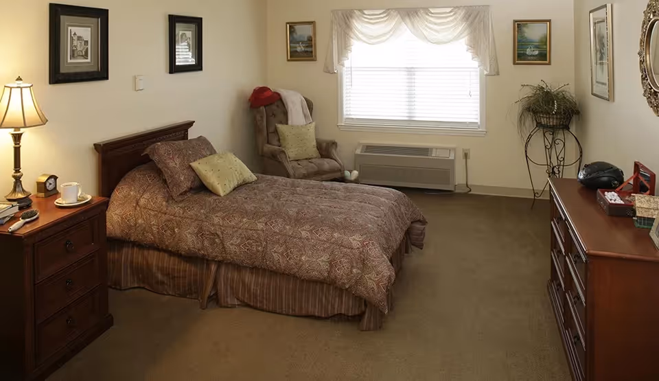 Cozy furnished bedroom with a double bed, bedside table and lamp, an armchair by a window, and a dresser.
