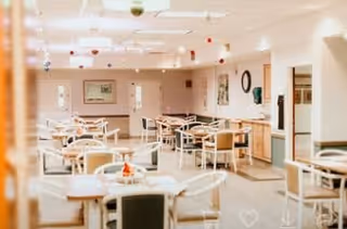 A spacious dining room in a senior living community with multiple tables and chairs arranged neatly. The room is well-lit with ceiling lights and decorated with small hanging ornaments. There are cabinets and a clock on the far wall, and the overall atmosphere is clean and welcoming.