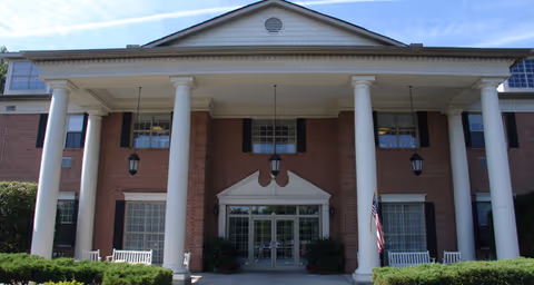 Front exterior view of a brick building with large white columns and a triangular pediment above the entrance. There are two white benches on either side of the entrance, hanging lanterns, and an American flag displayed near the right side. The sky is clear and blue.