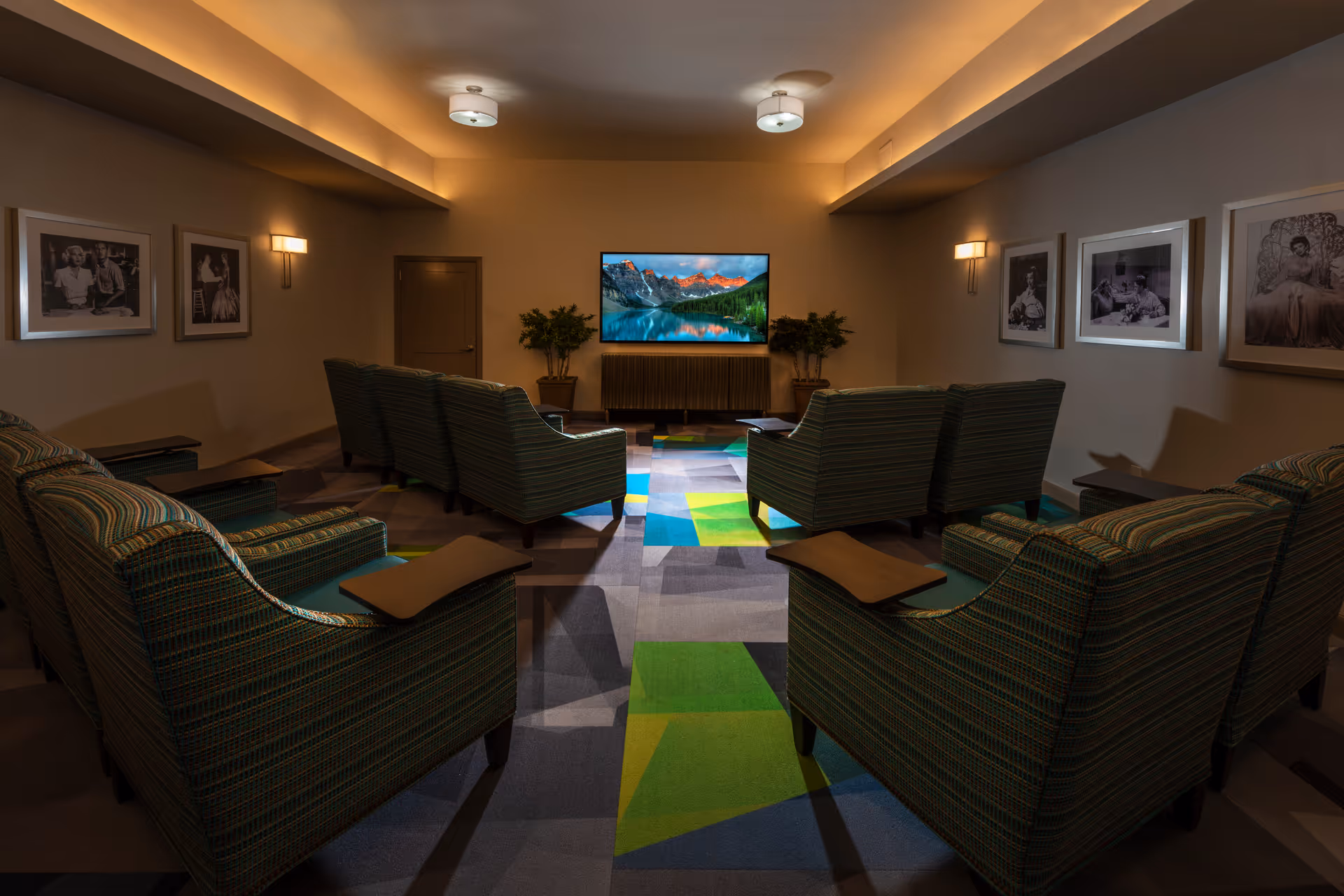 Dimly lit media room with rows of upholstered armchairs facing a wall-mounted TV and framed photos on the walls.