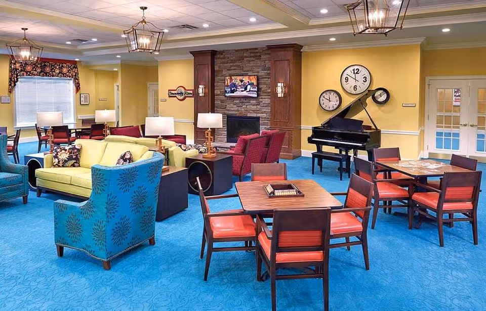 A spacious and brightly lit common area in an assisted living facility featuring blue carpet, yellow walls, and various seating options including green and blue armchairs, red cushioned chairs around wooden tables, and a green sofa. A black grand piano is positioned against the wall with three clocks above it. A stone fireplace with a mounted TV is in the background, and there are multiple lamps and ceiling lights providing illumination.