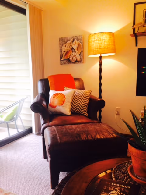 Cozy living room corner with a leather chaise lounge topped with decorative pillows, a floor lamp, and a sliding glass door to a balcony.