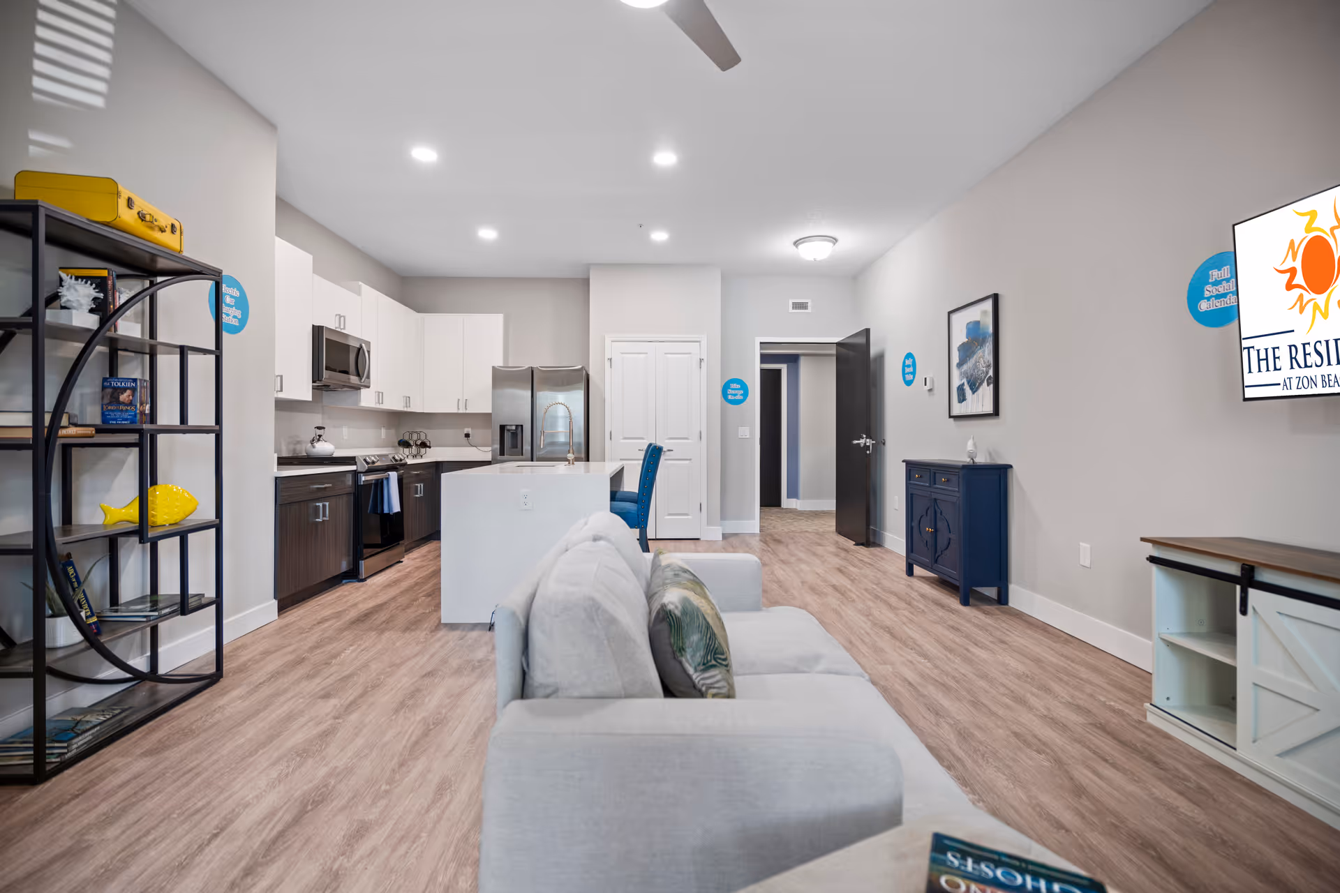 A modern open-concept living area featuring a light gray sofa with decorative pillows, a kitchen with white upper cabinets and dark lower cabinets, stainless steel appliances including a refrigerator and microwave, a kitchen island with a sink and blue bar stools, a black metal shelving unit with decorative items, wood flooring, and a wall-mounted TV displaying The Residences at Zon Beachside logo.