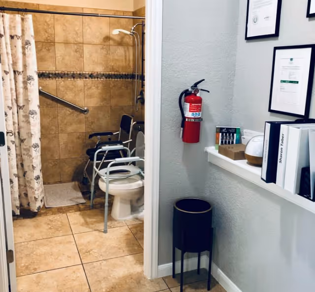 View into a bathroom with beige tiled walls and floor, featuring a shower area with a floral shower curtain, a grab bar, and a shower chair. In front of the shower is a toilet with a raised seat and armrests. Outside the bathroom, a wall-mounted fire extinguisher is visible along with a small shelf holding framed certificates, binders, and a diffuser. A black trash can is positioned below the shelf.