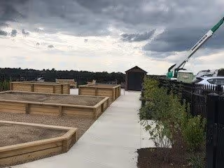 Outdoor raised garden beds with a concrete walkway between them, a small shed at the end of the path, a crane in the background, and a cloudy sky overhead.