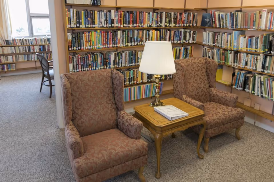 A cozy library corner with two patterned armchairs facing a wooden side table with a lamp and a book on it. Behind the chairs are shelves filled with books, and a window with curtains is visible in the background.