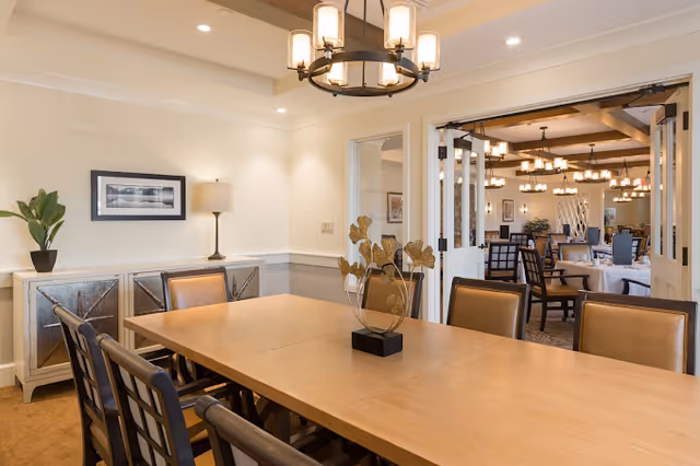 A well-lit dining room with a large wooden table surrounded by chairs. A decorative centerpiece with golden leaves is placed on the table. In the background, there is a sideboard with a potted plant and a lamp, and a framed picture on the wall. Through open double doors, another dining area with multiple tables and chairs is visible, featuring similar lighting fixtures and decor.