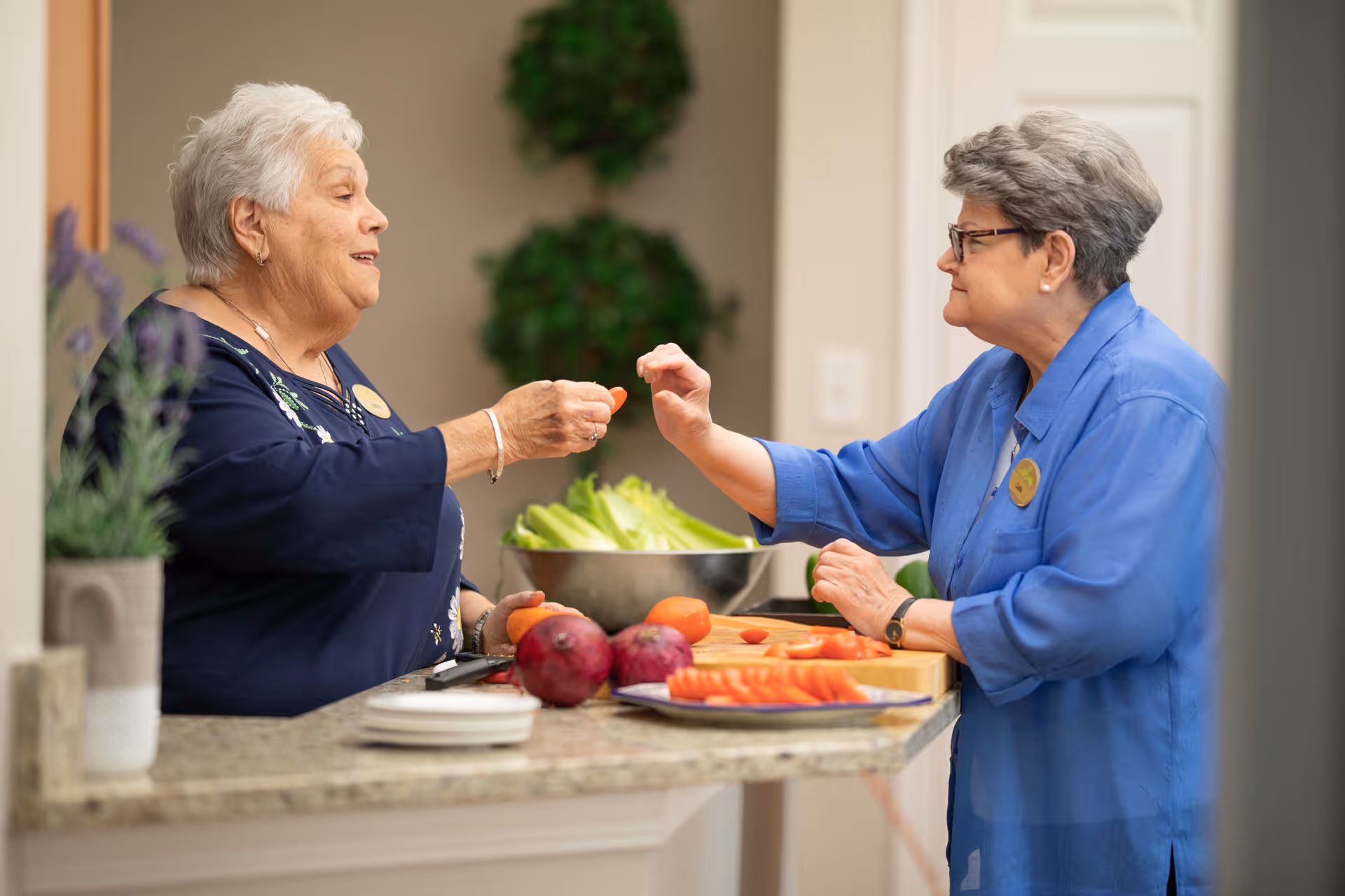 Two elderly women standing at a kitchen counter, one handing a piece of sliced carrot to the other. The counter has a cutting board with sliced carrots, whole carrots, and onions, with a bowl of lettuce in the background.