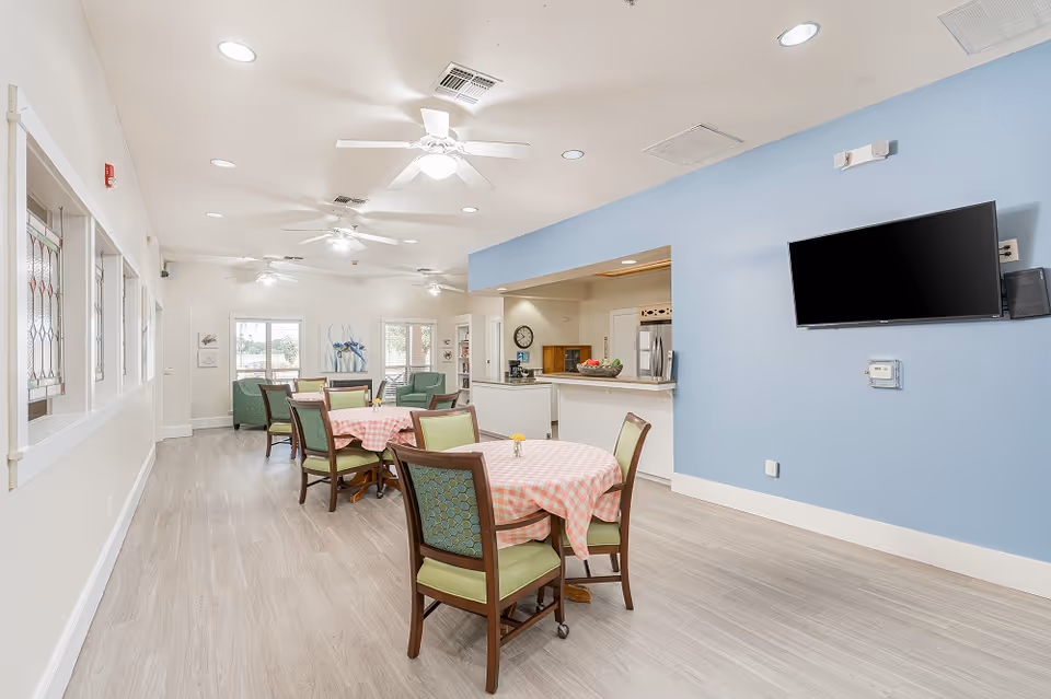 A bright and spacious common area in an assisted living facility featuring several round tables with pink checkered tablecloths and green cushioned chairs. The room has light wood flooring, white walls with a blue accent wall, ceiling fans, and recessed lighting. A flat-screen TV is mounted on the blue wall, and a kitchen area with a counter and stainless steel appliances is visible in the background. Large windows and glass doors allow natural light to enter the space.