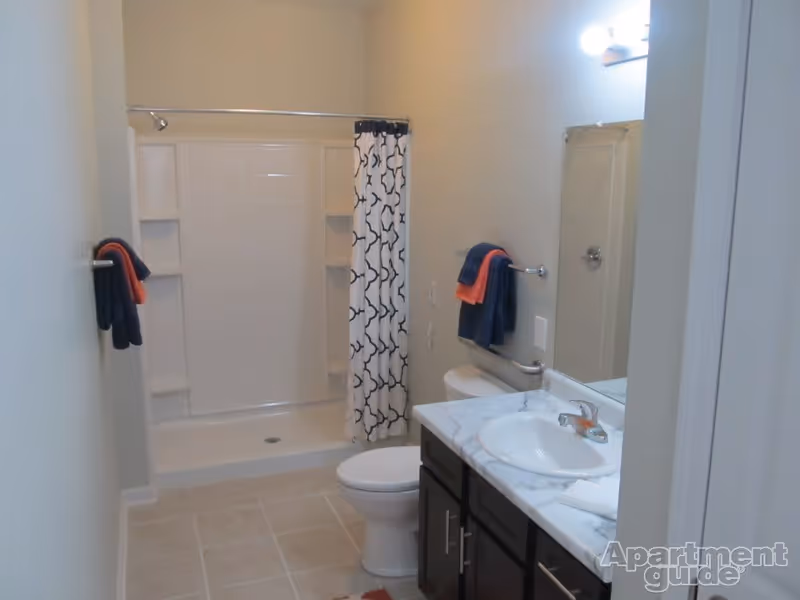 A clean bathroom with a white toilet, a sink with a marble countertop, a large mirror, and a shower with a white and black patterned curtain. There are towels hanging on the wall, and the floor is tiled.