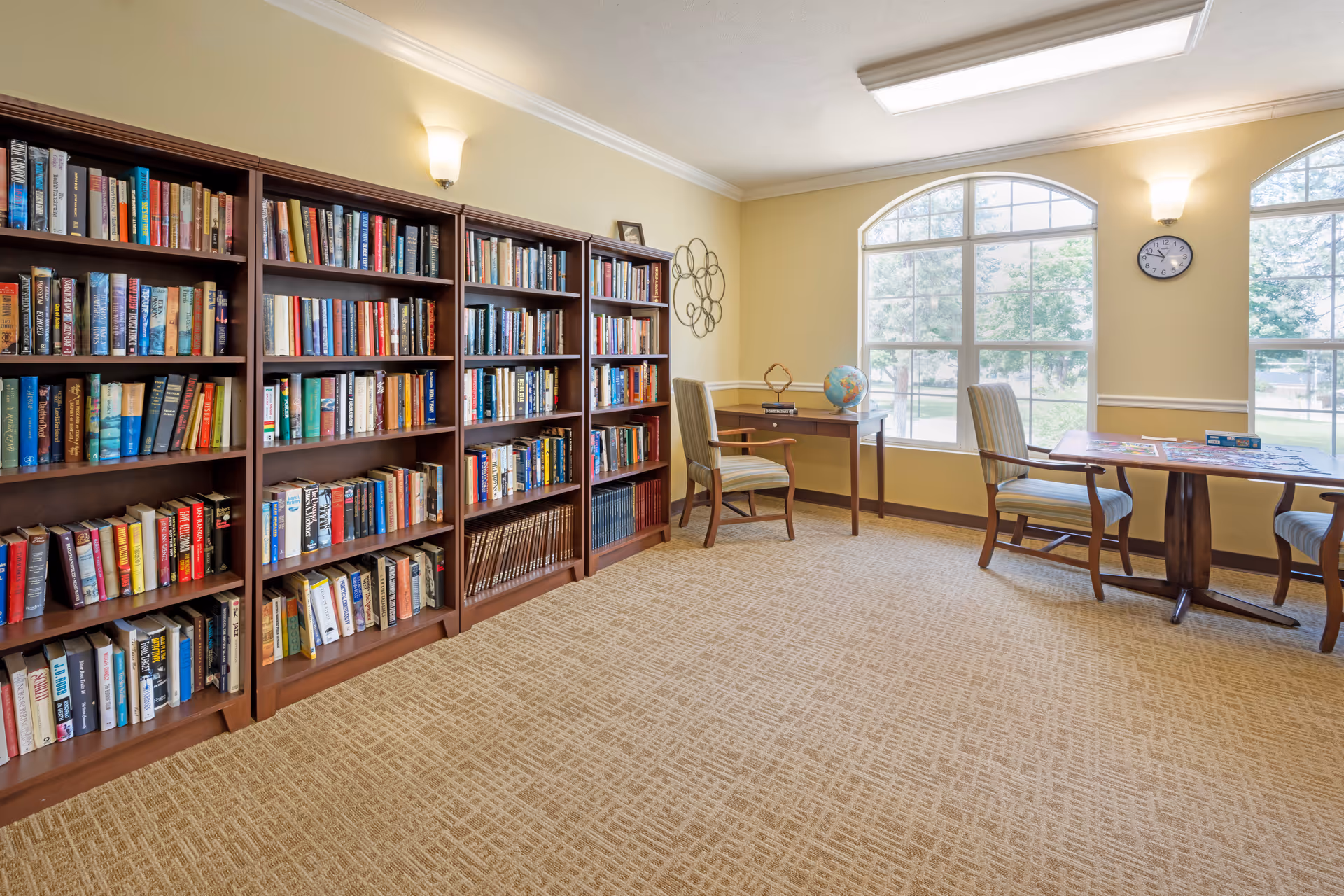 A bright and cozy library room with large windows letting in natural light. The room features several wooden bookshelves filled with books along one wall, two wooden tables with chairs, a globe, and a wall clock. The walls are painted light yellow and the floor is carpeted in a beige pattern.