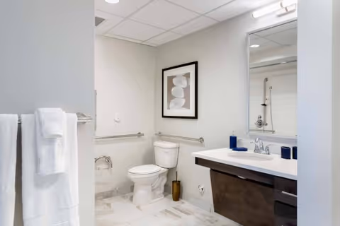 A clean and modern bathroom with a white toilet, a large mirror above a dark wood vanity with a white countertop, and a towel rack holding white towels. The walls are light-colored, and there is a framed abstract artwork above the toilet. The floor is tiled in a light pattern.