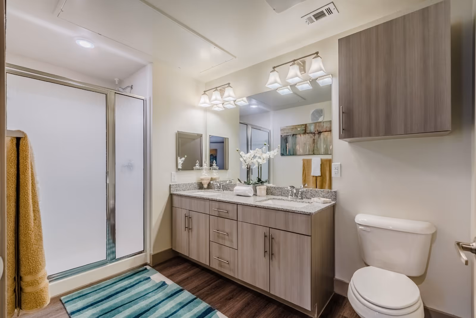 A modern bathroom featuring a double sink vanity with granite countertop, two mirrors, and overhead lighting. There is a white toilet next to the vanity and a shower with frosted glass sliding doors. The floor has a striped blue and green rug, and yellow towels hang on a rack. A wooden cabinet is mounted on the wall above the toilet.