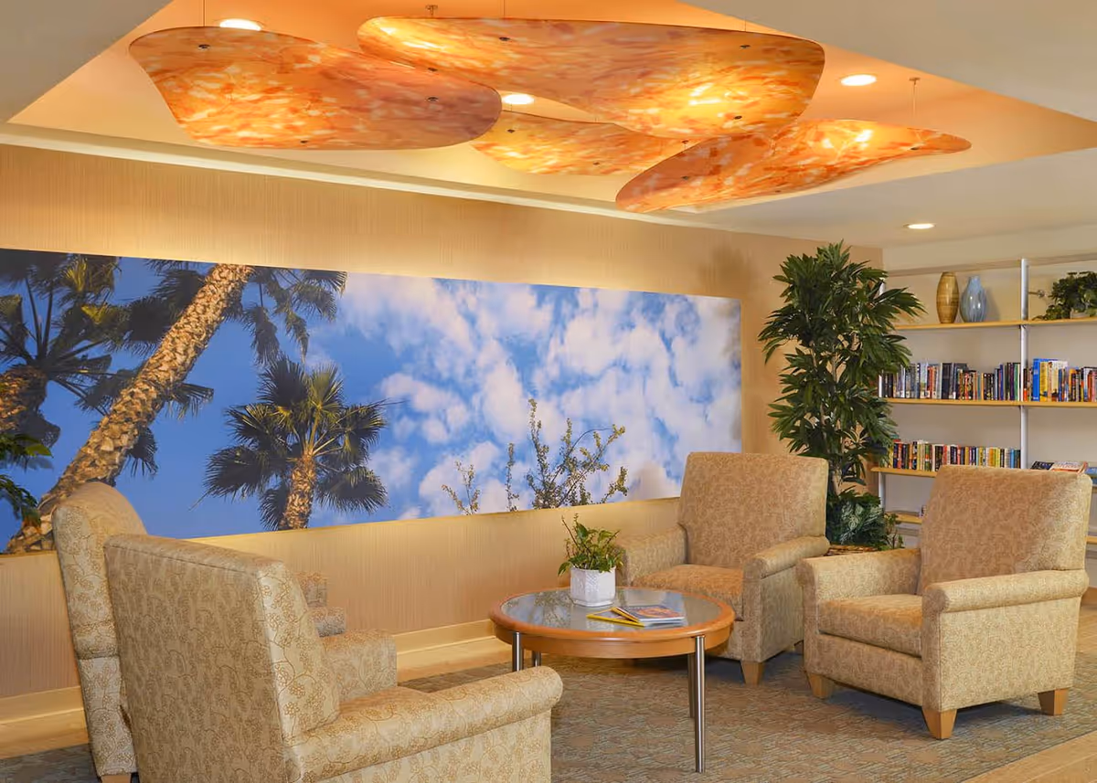 A cozy sitting area with four beige patterned armchairs arranged around a round wooden coffee table with a small plant and magazines on it. Behind the chairs is a large wall art featuring a blue sky with clouds and palm trees. To the right, there is a tall green plant and a bookshelf filled with books and decorative items. The ceiling has an artistic orange and yellow light fixture.