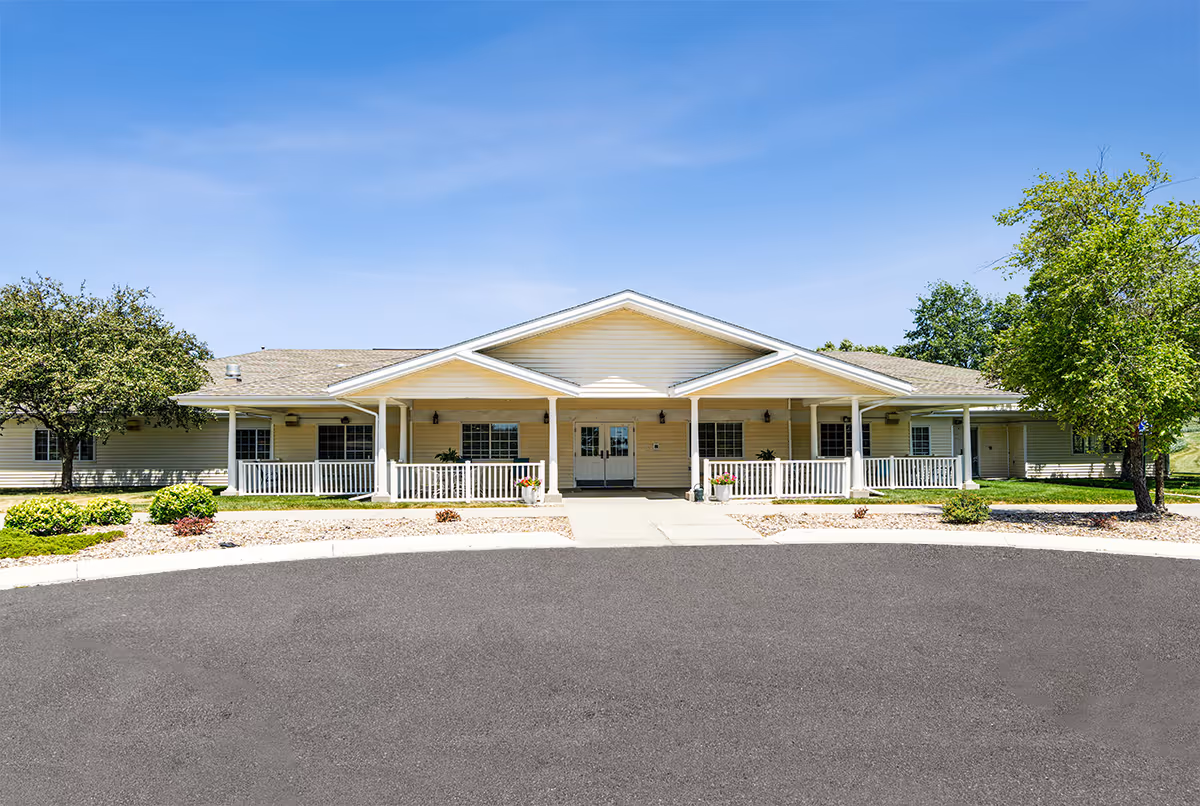 Front exterior view of a single-story building with a covered porch, white railings, and a paved driveway in front. The building is surrounded by green trees and shrubs under a clear blue sky.