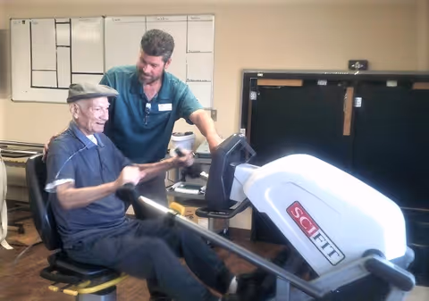An elderly man wearing a cap is using a SCIFIT rowing exercise machine while a male staff member in a green shirt assists and encourages him in a room with beige walls and a whiteboard in the background.
