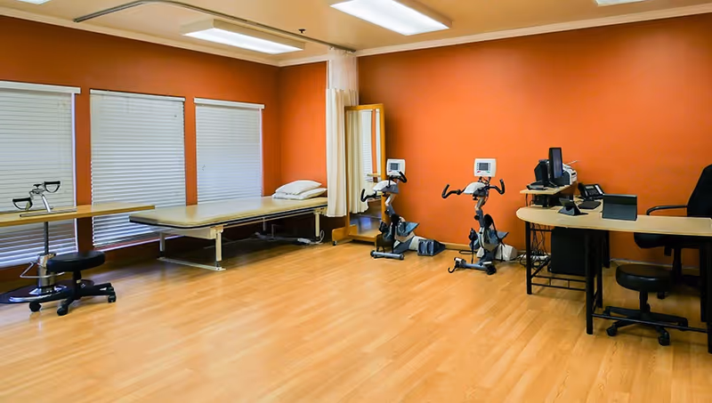 Spacious therapy room with stationary exercise bikes, a treatment table, desks and wood flooring against an orange wall.