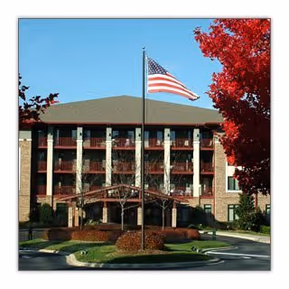 Exterior view of a multi-story senior living facility building with a flagpole flying the American flag in front. The building has balconies and is surrounded by landscaped greenery and trees with red leaves.