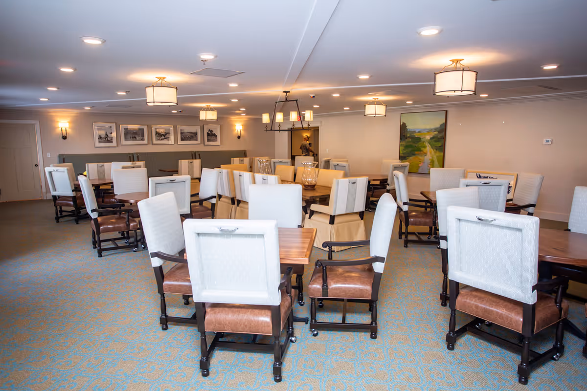 A spacious dining room with multiple wooden tables and cushioned chairs arranged neatly on a patterned carpet. The room is well-lit with ceiling lights and wall sconces, and decorated with framed pictures and a large painting on the walls.