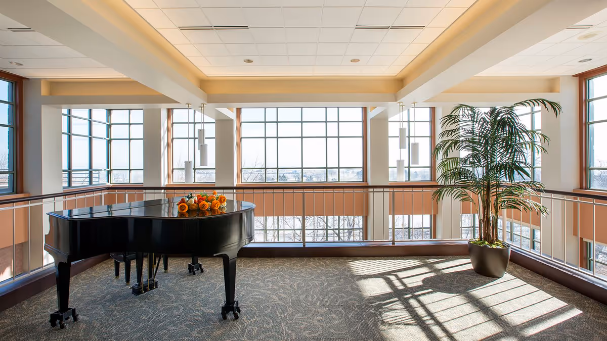 A bright and spacious interior area with large windows letting in natural light. A black grand piano decorated with a floral arrangement is positioned on the left side, and a tall potted plant is on the right. The space has carpeted flooring and modern ceiling lights.