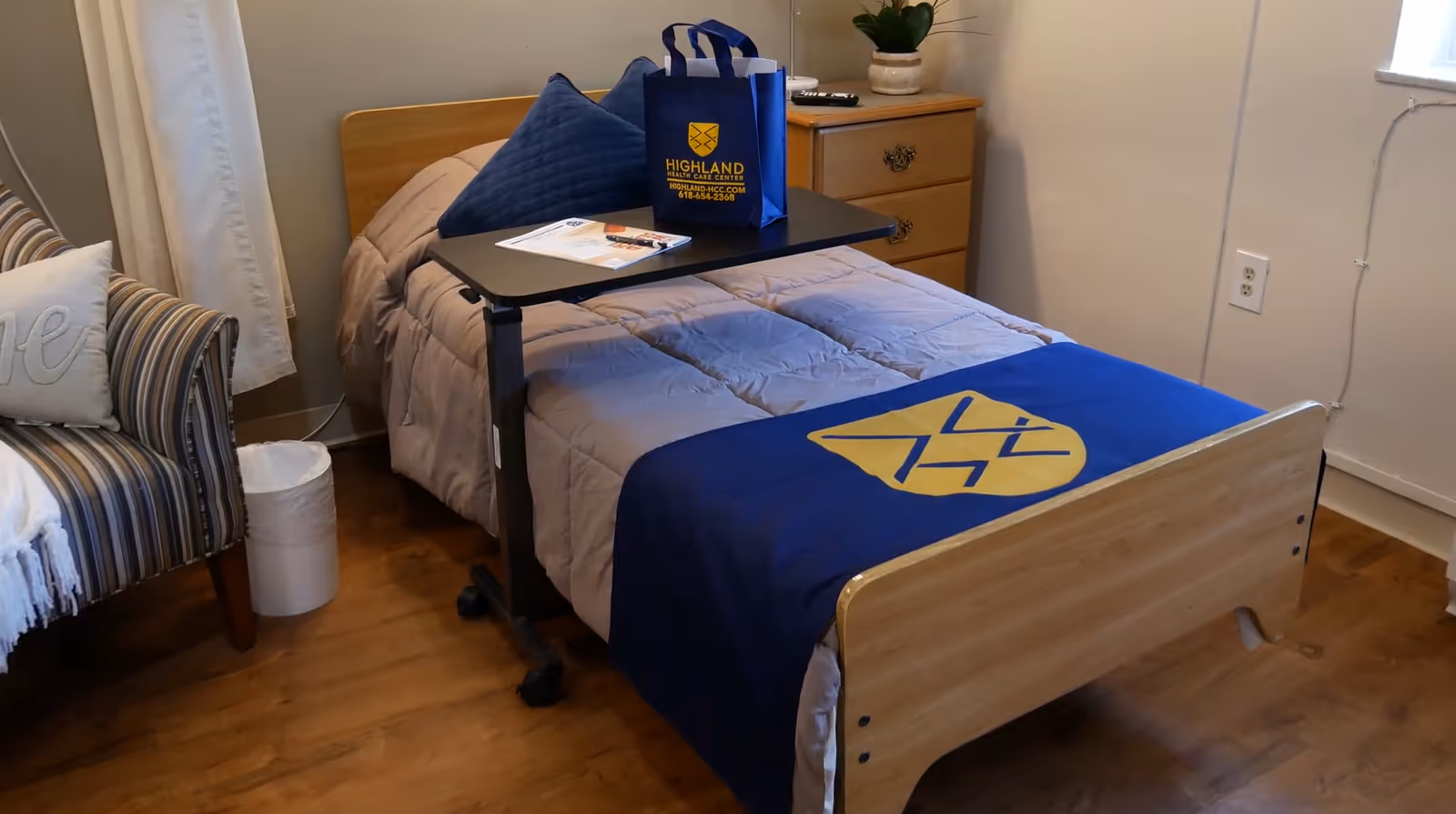 A single bed with a wooden headboard and footboard in a small room. The bed is made with a beige comforter and a blue blanket featuring a yellow emblem. On the bed is a black overbed table holding a blue gift bag with the Highland Healthcare Center logo and some papers. Next to the bed is a wooden dresser with a potted plant on top. To the left of the bed is a striped armchair with a white pillow and a white throw blanket. The room has wooden flooring and light-colored walls with a window partially covered by white curtains.