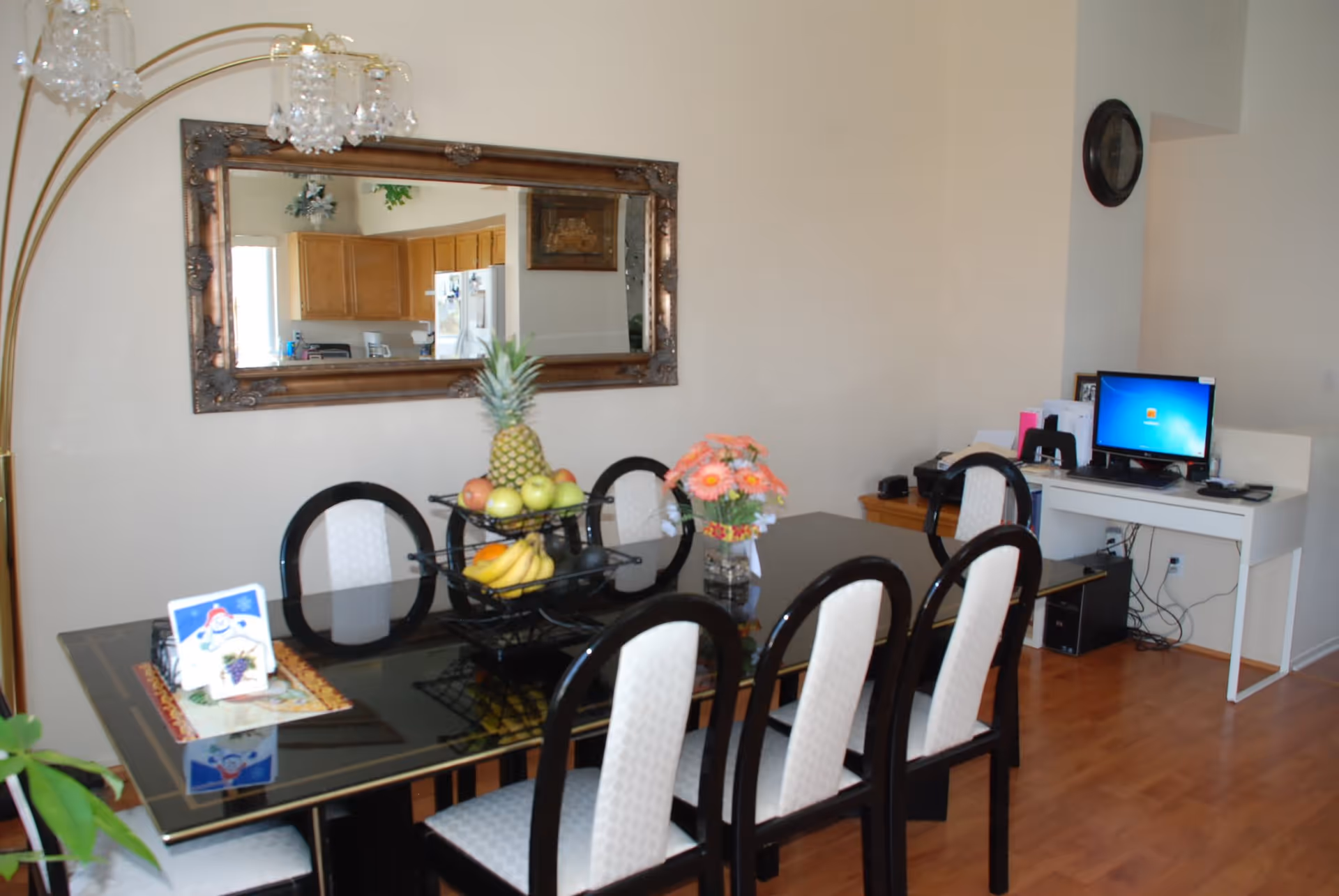 Dining room with a black glass table surrounded by six chairs with white cushions. On the table, there is a two-tiered fruit basket containing a pineapple, apples, bananas, and other fruits, along with a vase of pink flowers and a small decorative card. A large ornate mirror hangs on the wall reflecting a kitchen area. To the right, there is a white desk with a computer and various office supplies. The floor is wooden, and a tall lamp with crystal-like shades stands to the left.