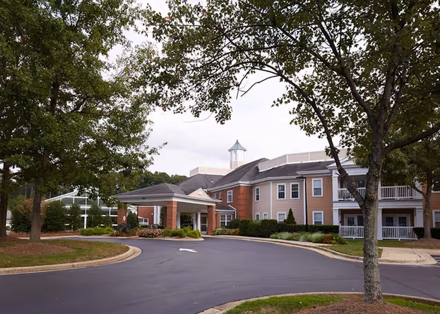 Exterior view of a senior living facility named Woodland Terrace, showing a two-story building with beige and brick siding, a covered entrance, and surrounding trees and landscaping.