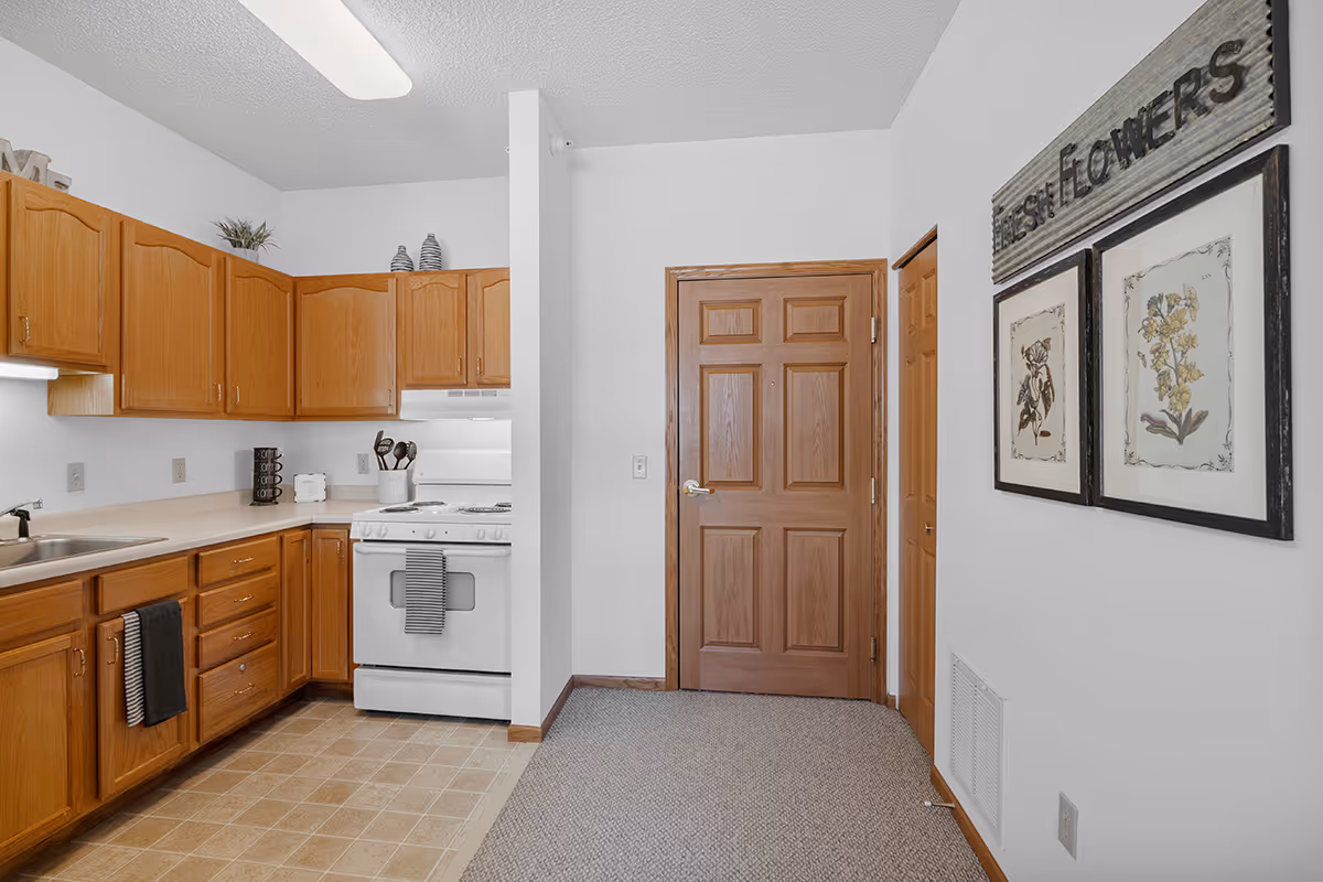 A kitchen area with wooden cabinets, a white stove with a striped towel hanging on the handle, a sink with a black towel hanging from the cabinet, and a countertop with kitchen utensils and decor. There is a wooden door and two framed botanical prints on the adjacent wall with a sign above them that reads 'Fresh Flowers'.