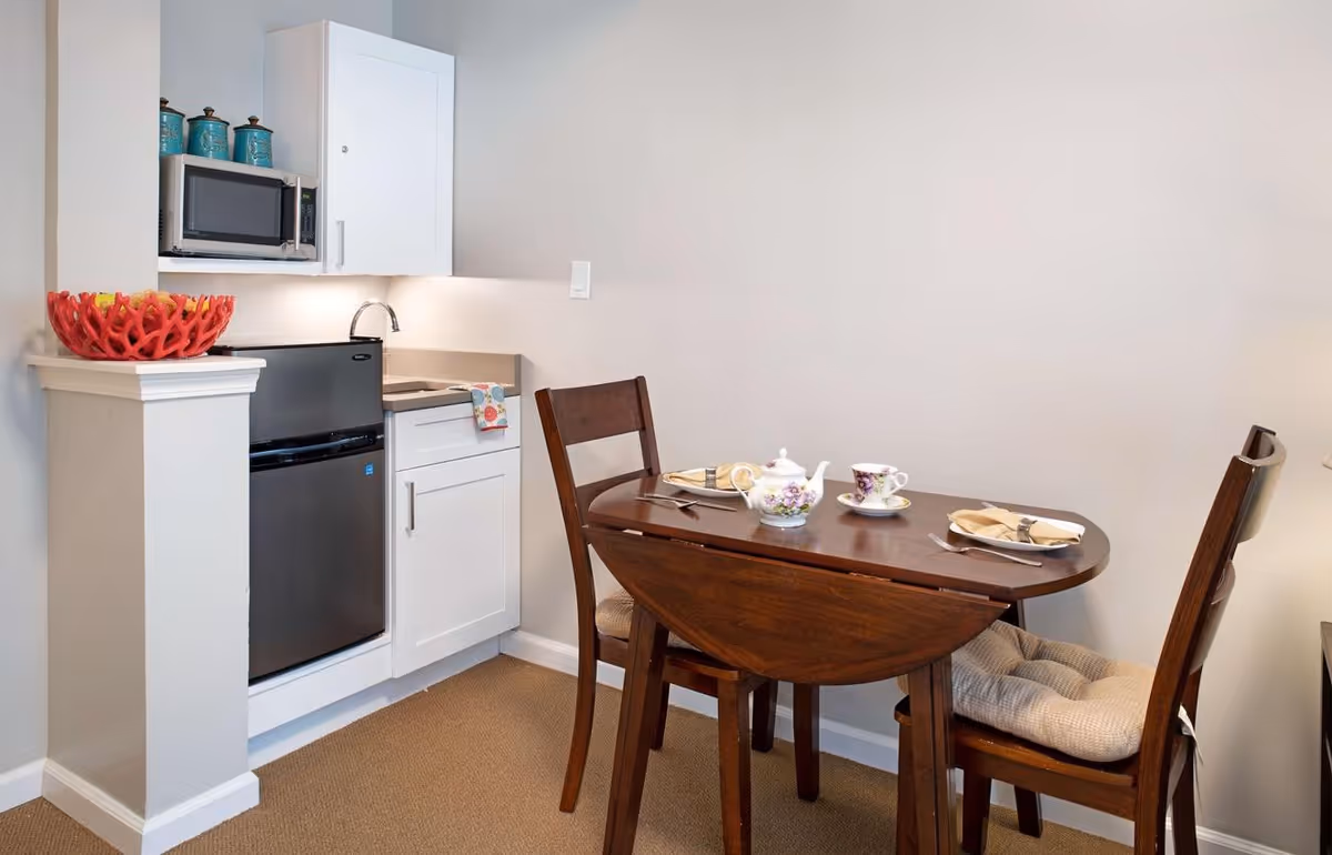 Small kitchenette area with a mini refrigerator, microwave, sink, and white cabinets. Next to the kitchenette is a wooden dining table set for tea with two chairs, each with a cushion. The table has a teapot, teacups, and plates with napkins and cutlery.