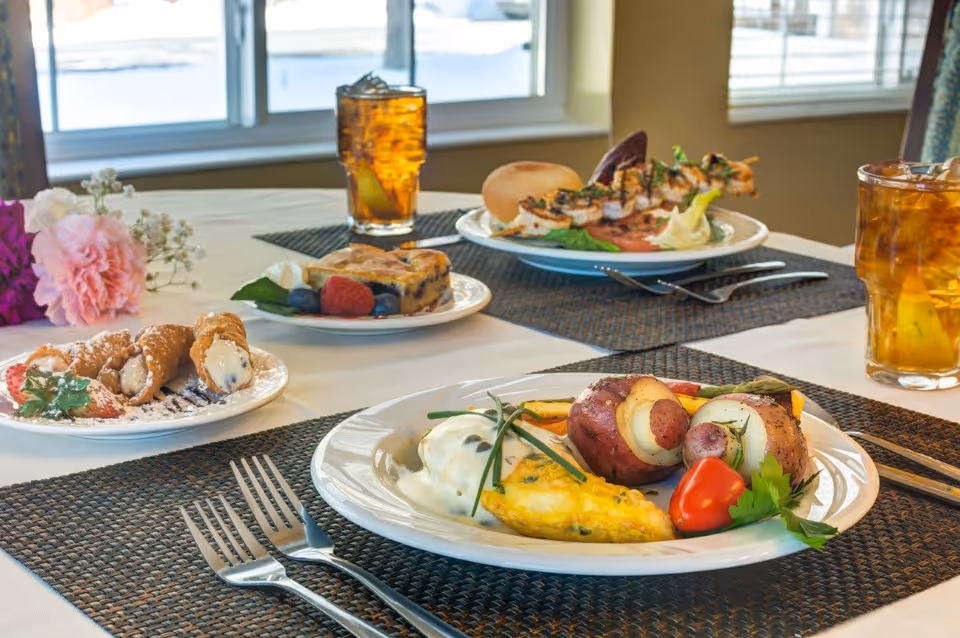 A dining table set with plates of food including an omelette with roasted potatoes and vegetables, a dessert plate with cannoli and strawberries, a slice of berry pie with fresh berries, and a plate with skewered shrimp and a bread roll. Two glasses of iced tea are also on the table, which is covered with a white tablecloth and dark placemats. A window with blinds is visible in the background.