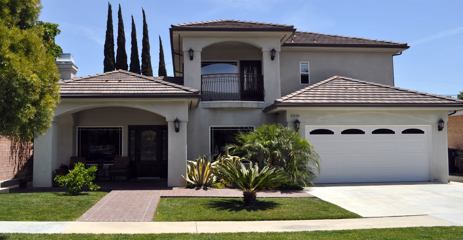 Front exterior view of a two-story residential building with a tiled roof, a balcony with wrought iron railing, a double garage door, and a small landscaped front yard with green grass, shrubs, and trees.