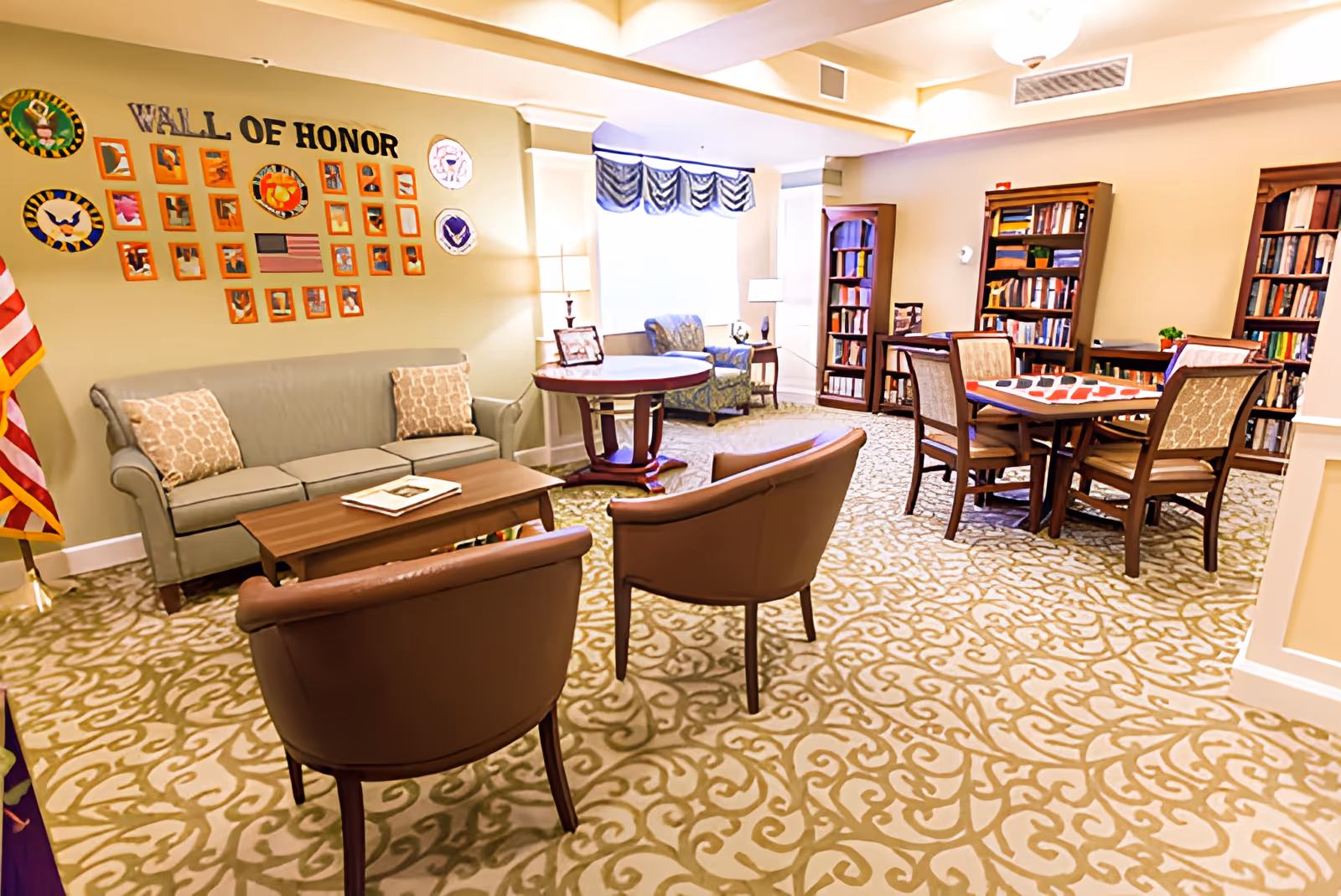 A cozy common area in a senior living facility featuring a 'Wall of Honor' with framed photos and plaques on the wall. The room includes a gray sofa with two patterned pillows, two brown armchairs, a wooden coffee table, a round side table with a lamp, and a blue patterned armchair near a window with a valance. There are also bookshelves filled with books and a table with four chairs, some with a checkered board game on top. The carpet has an intricate beige and green pattern.