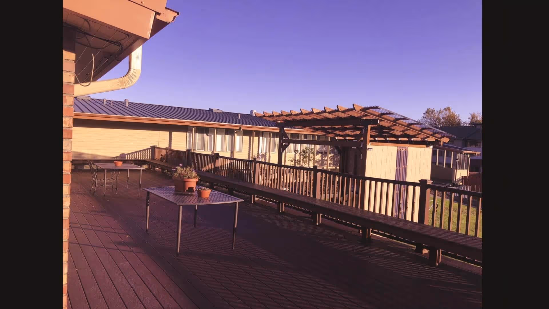 Outdoor wooden deck area with metal tables and potted plants, a wooden pergola, railing, and a building with windows in the background under a clear blue sky.