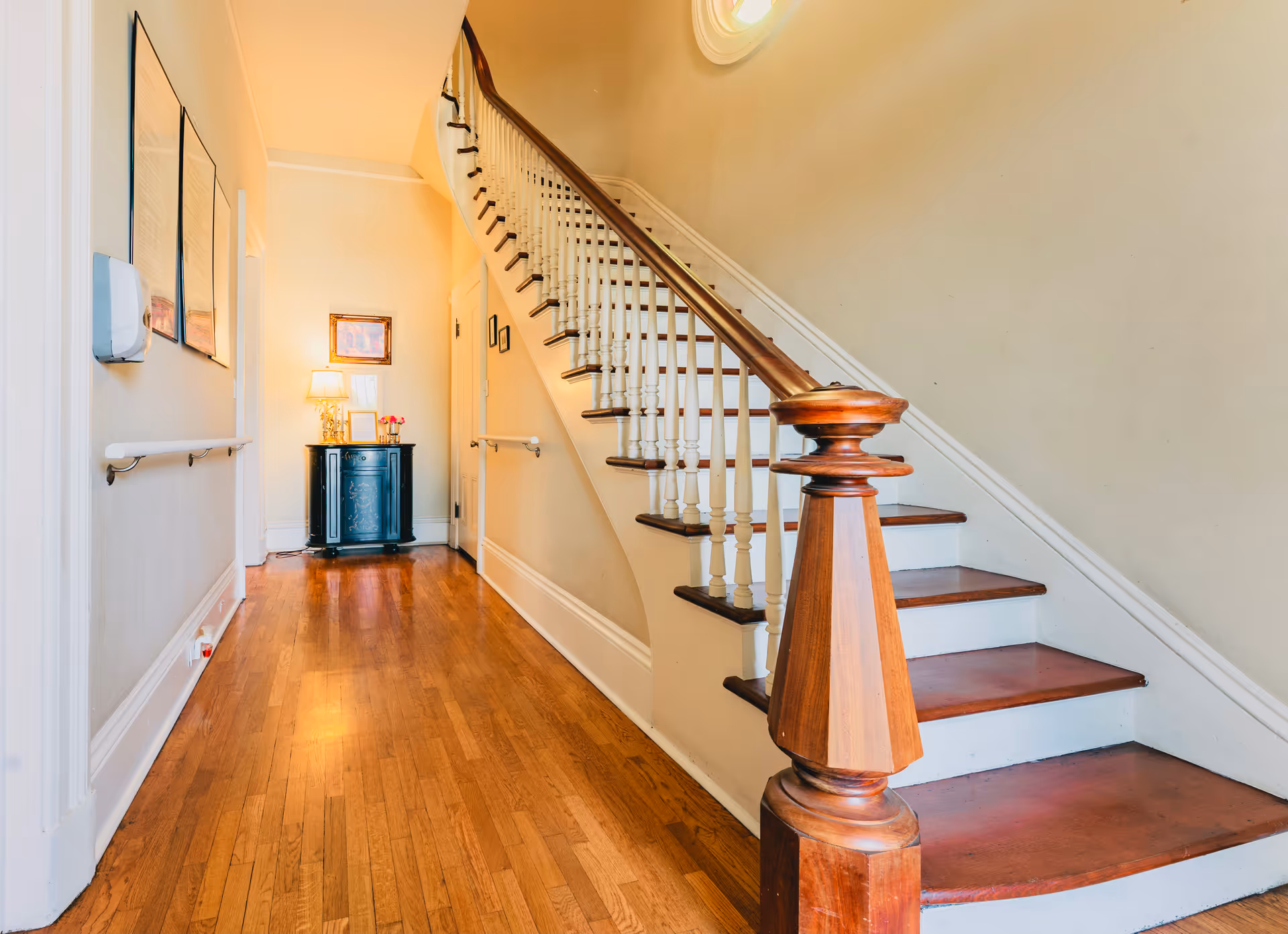 A hallway with wooden floors and a staircase leading upward, featuring a lamp and decor along the walls.