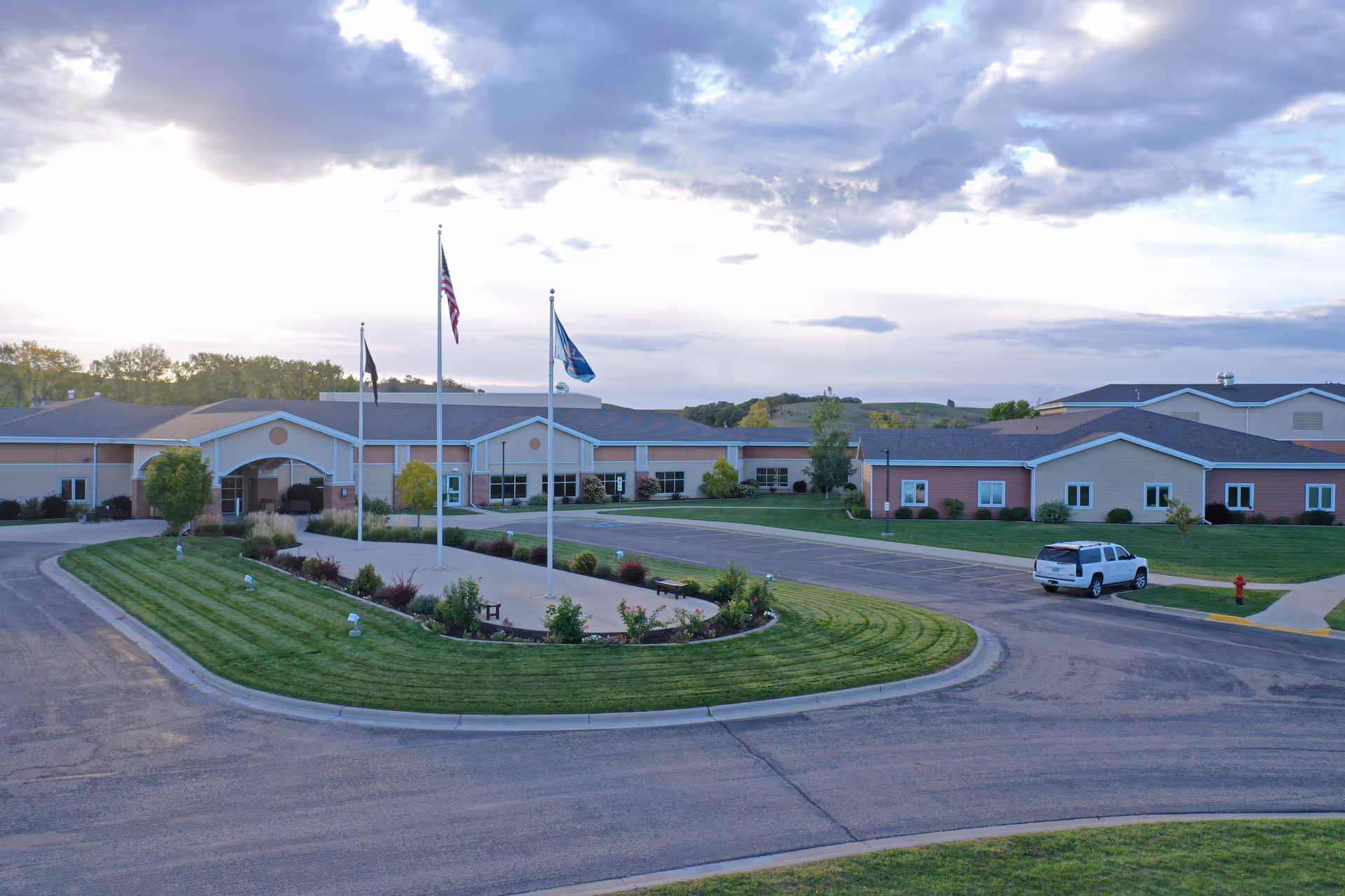 Exterior view of the North Dakota Veterans Home building with a circular driveway, three flagpoles with flags, well-maintained landscaping, and a white SUV parked near the entrance under a partly cloudy sky.