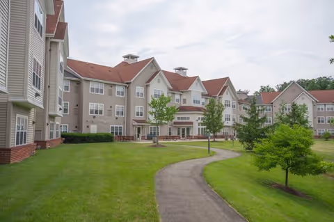 Exterior view of a senior living facility with beige buildings featuring red roofs and multiple windows. A paved walkway curves through a well-maintained green lawn with small trees and shrubs under a cloudy sky.