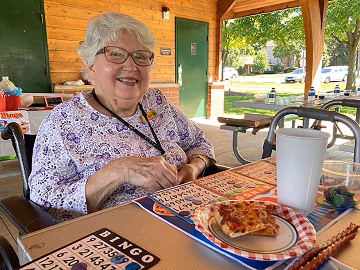 An elderly woman with white hair and glasses sitting at a picnic table under a wooden pavilion. She is smiling and playing bingo with several bingo cards in front of her. On the table, there is a paper plate with a slice of pizza, a large white cup, and a small clear cup with marbles. The background shows green grass, trees, and parked cars.
