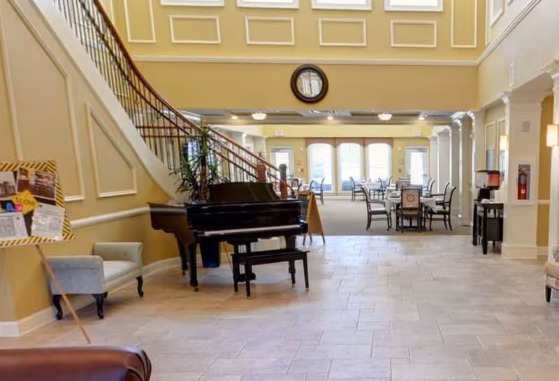 Spacious senior living facility common area with a grand piano and bench near a staircase, several chairs and tables in the background, beige walls with decorative molding, and large windows letting in natural light.