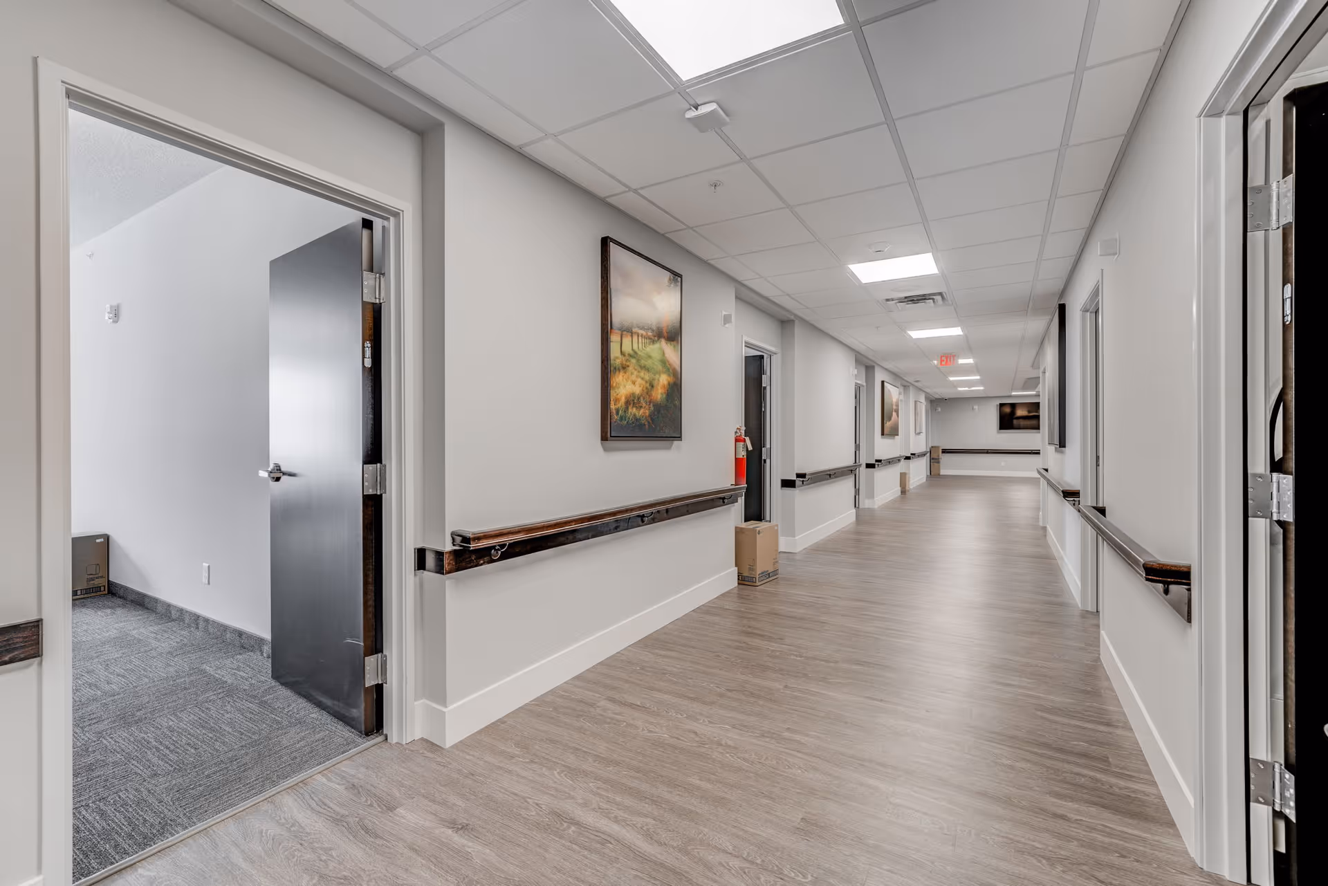 Long, well-lit interior corridor of a senior living facility with handrails, open doors, and framed artwork on the walls.