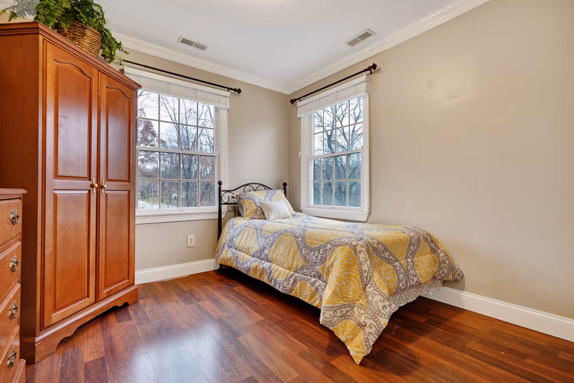 A small bedroom with two large windows letting in natural light. The room features a single bed with a yellow and gray patterned comforter and pillows. There is a wooden armoire and a matching dresser with a plant on top. The floor is hardwood and the walls are painted beige.