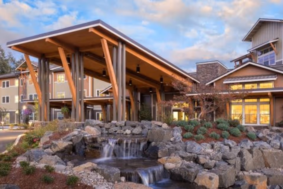 Exterior view of The Springs at Greer Gardens facility featuring a large covered entrance with wooden beams, a landscaped rock waterfall in the foreground, and a multi-story building with large windows illuminated from inside under a partly cloudy sky.