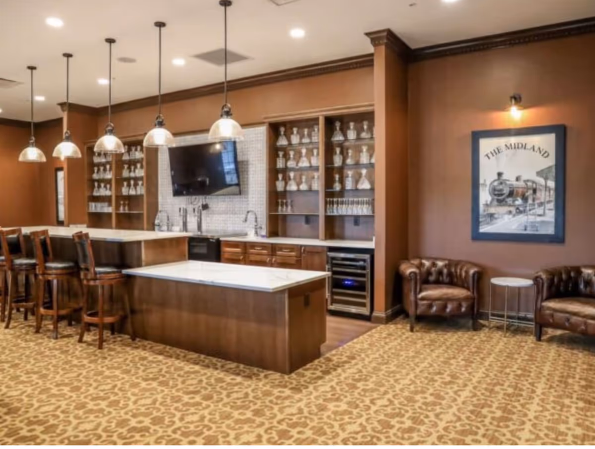 Communal bar and lounge area with a marble-topped island, bar stools, pendant lights, shelving of glassware, a wall-mounted TV, and leather armchairs.
