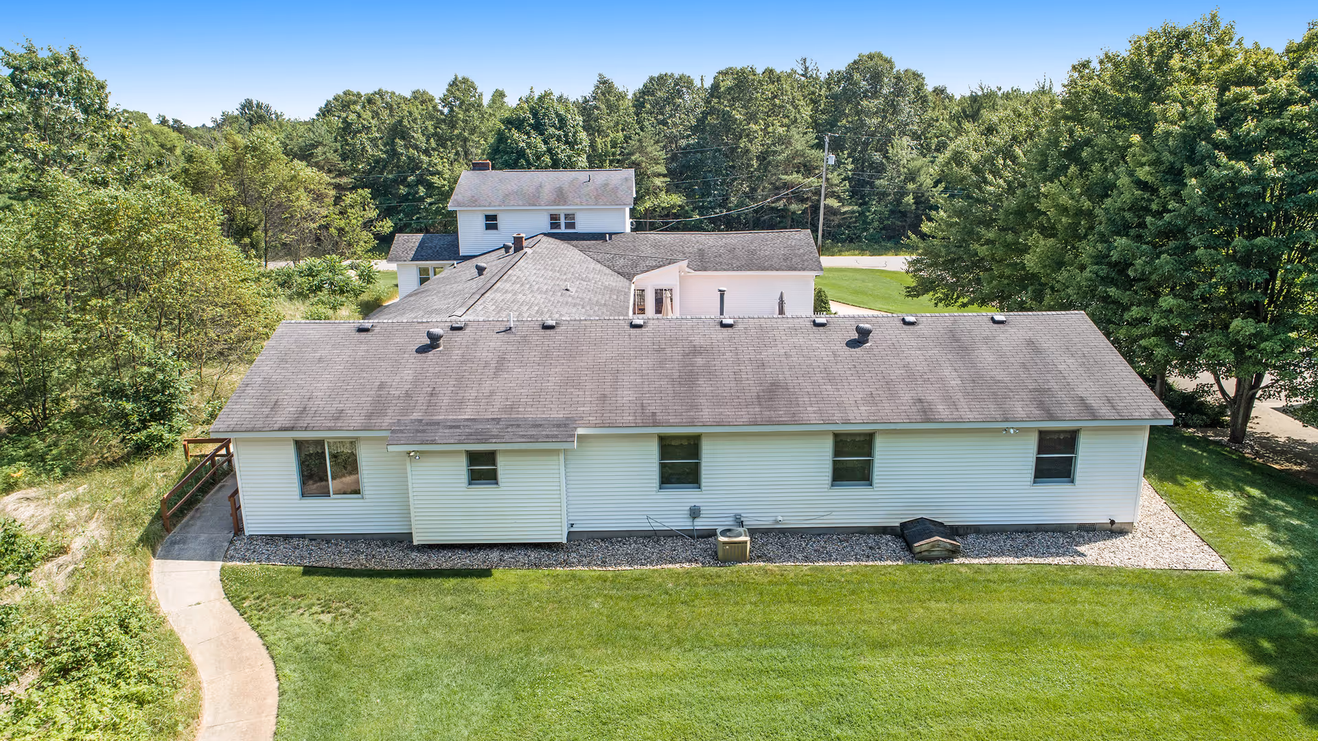 Aerial view of a single-story white building with a gray roof surrounded by green grass and trees under a clear blue sky.