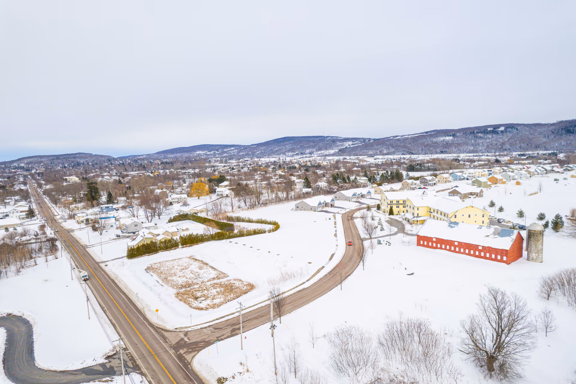 Aerial view of a snowy rural senior living campus with yellow buildings, a red barn and surrounding roads and houses.