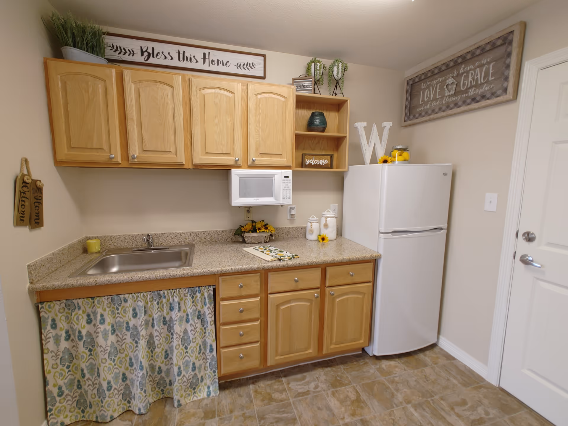Small kitchenette with light wood cabinets, a sink and microwave over a speckled countertop, and a white refrigerator.