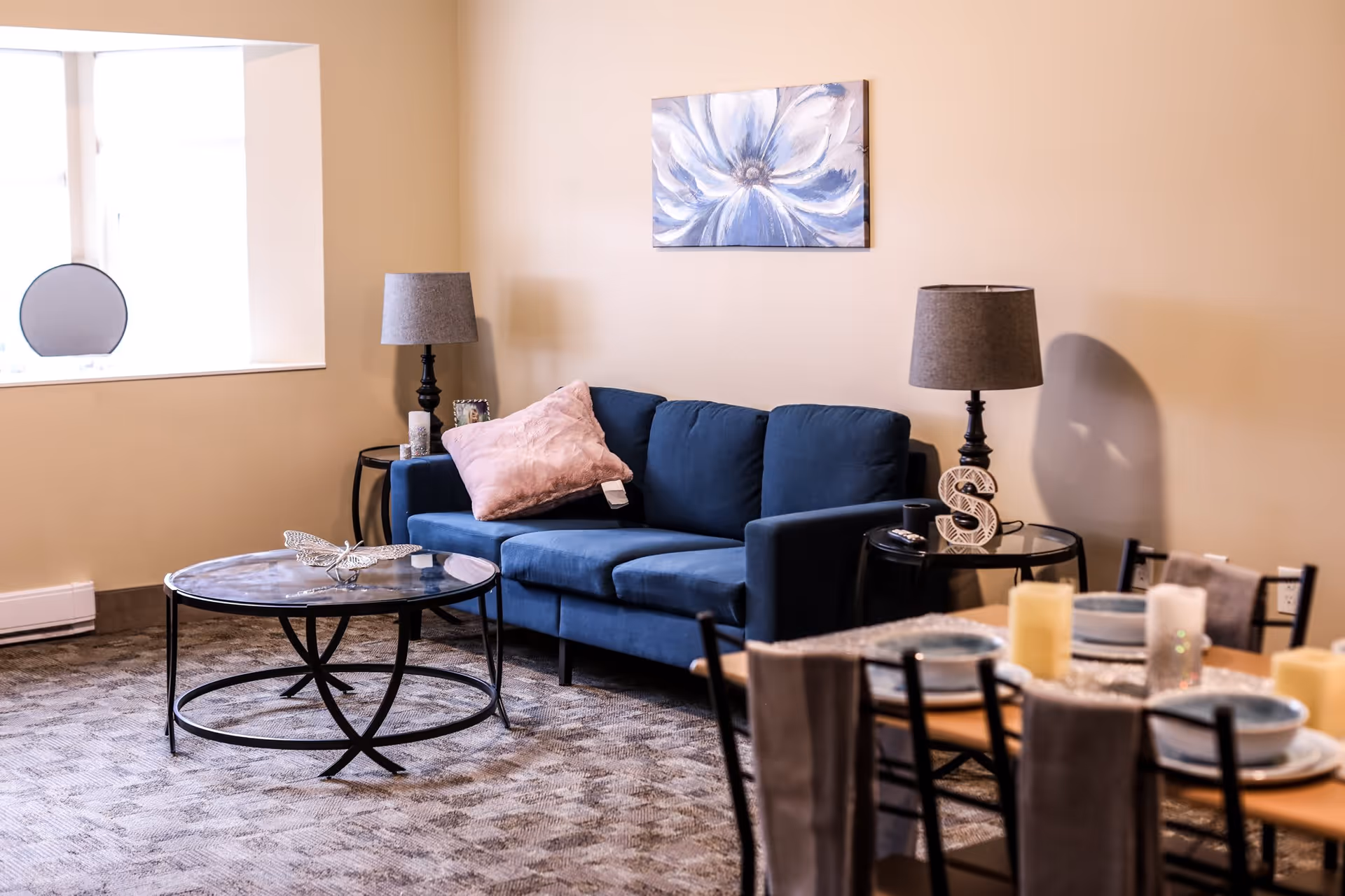 A cozy living room area with a blue sofa adorned with a pink pillow, two side tables each with a lamp, a round glass coffee table with a decorative butterfly, a dining table set with plates and candles, and a large window letting in natural light. A painting of a blue and white flower hangs on the beige wall behind the sofa.