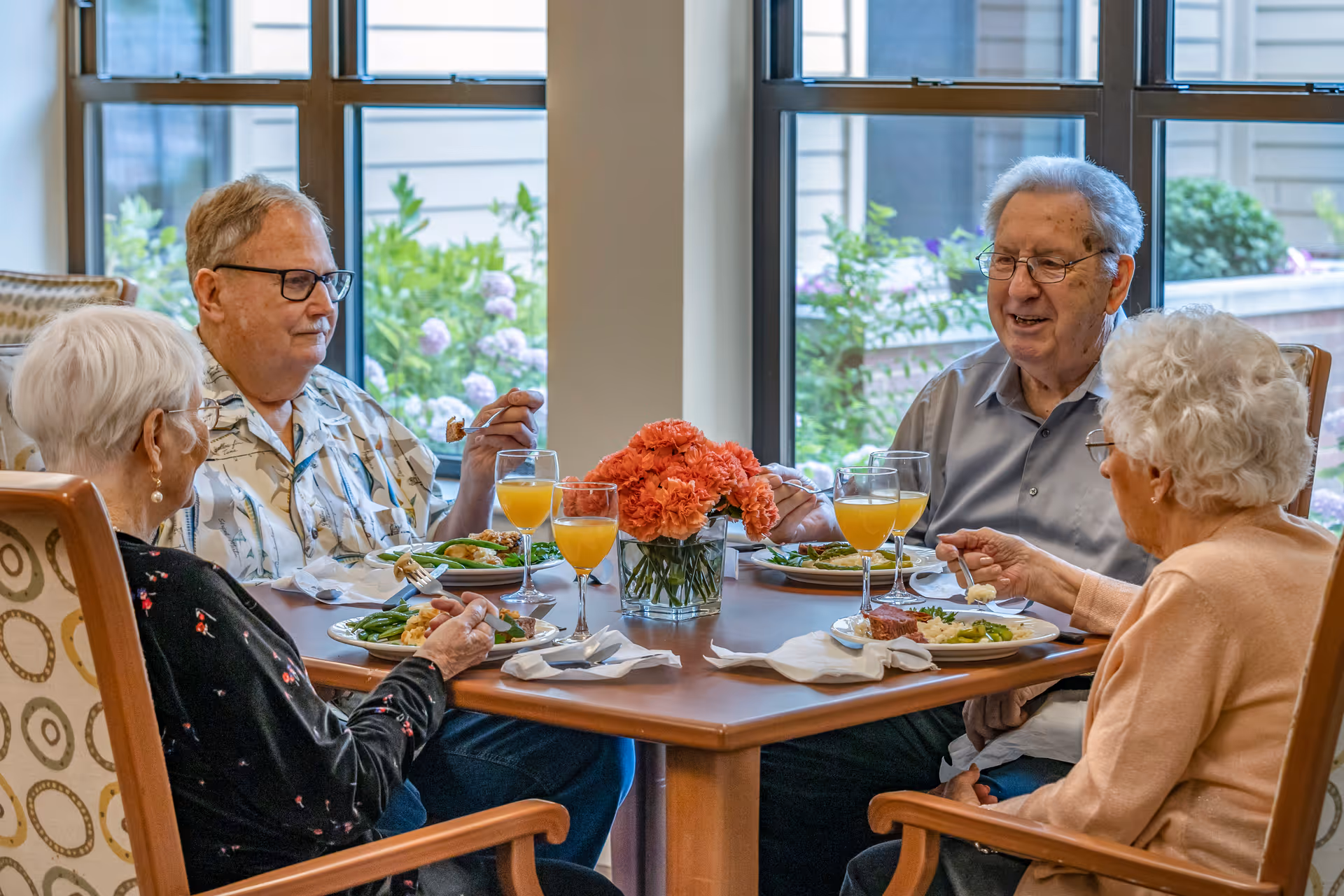 Four elderly residents sit around a table in a bright dining room sharing a meal with glasses of orange juice and a vase of flowers.