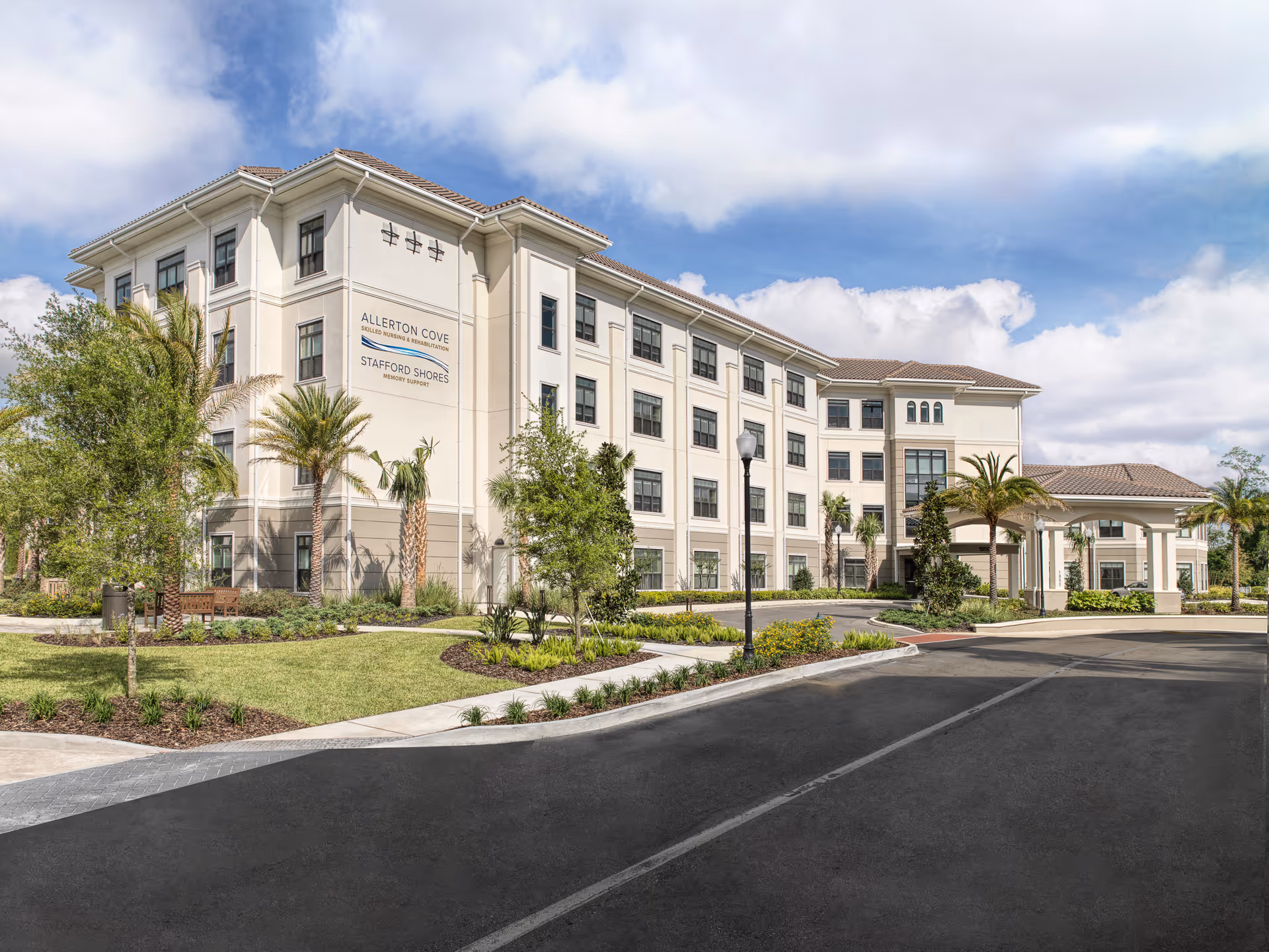Exterior view of a large, multi-story senior living facility building with beige walls and a tiled roof. The building is surrounded by landscaped greenery, palm trees, and a paved driveway under a partly cloudy sky.